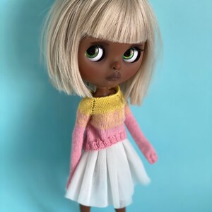 May include: A doll with black skin, white hair, and green eyes is wearing a pink and yellow striped sweater and a white skirt. The doll is standing on a light blue background.