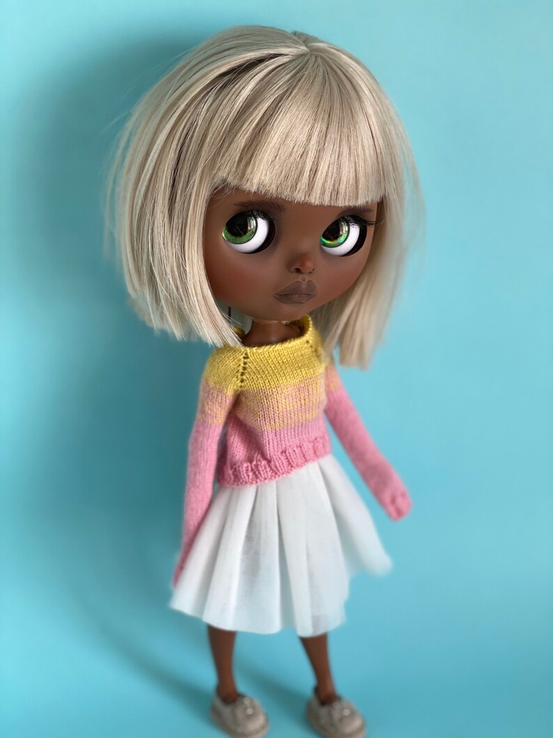 May include: A doll with black skin, white hair, and green eyes is wearing a pink and yellow striped sweater and a white skirt. The doll is standing on a light blue background.