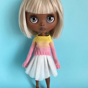 May include: A doll with blonde hair, brown skin, and blue eyes is wearing a pink and yellow sweater, a white tulle skirt, and brown shoes.