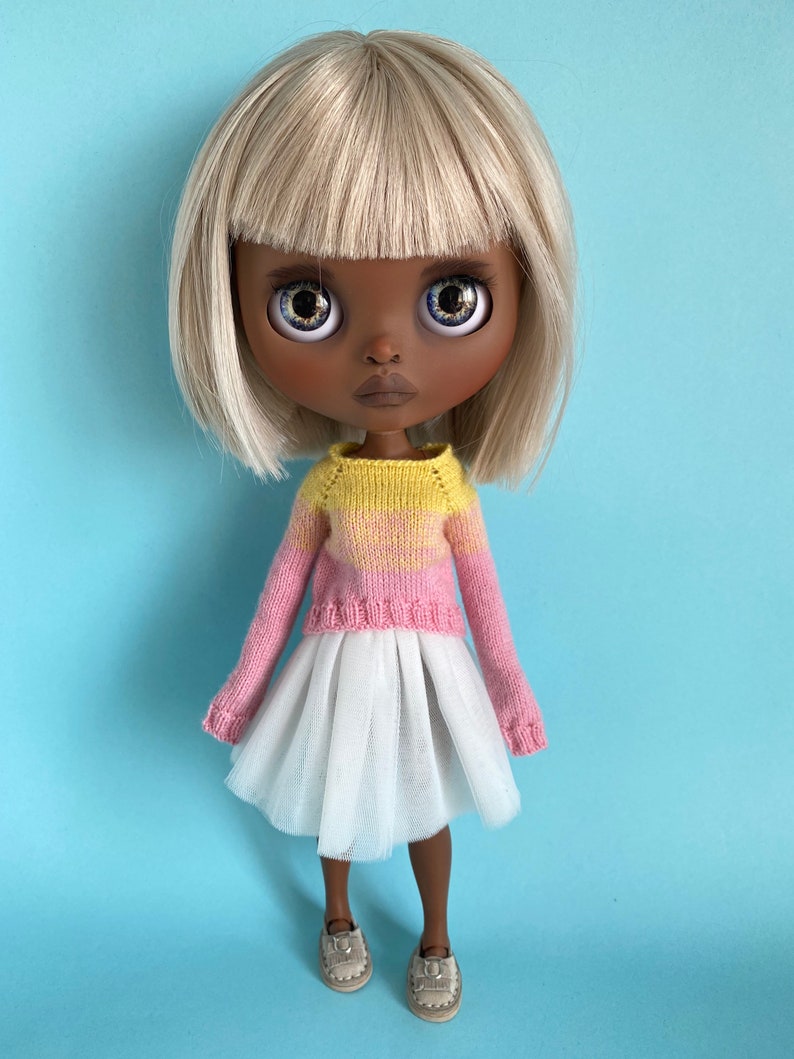 May include: A doll with blonde hair, brown skin, and blue eyes is wearing a pink and yellow sweater, a white tulle skirt, and brown shoes.