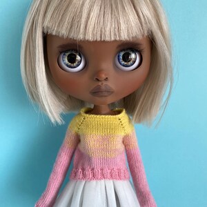 May include: A doll with blonde hair and brown skin wearing a pink and yellow striped sweater and a white tulle skirt.