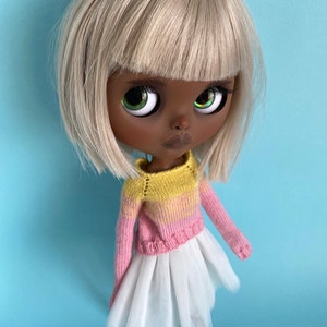 May include: A doll with black skin, white hair, and green eyes is wearing a pink and yellow striped sweater and a white tulle skirt.
