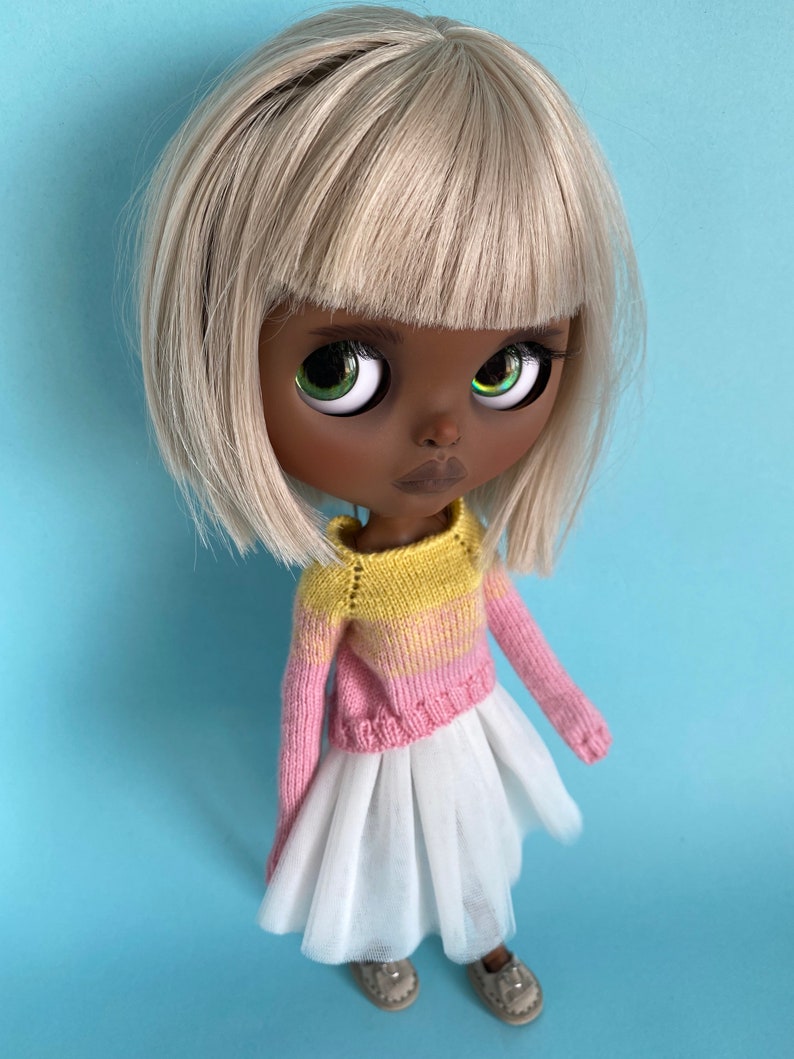 May include: A doll with black skin, white hair, and green eyes is wearing a pink and yellow striped sweater and a white tulle skirt.