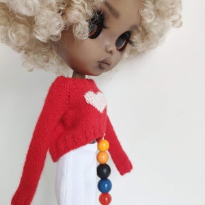 May include: A doll with brown skin and blonde curly hair is wearing a red sweater with a white heart on the front and white pants. The doll is wearing a colorful beaded necklace.