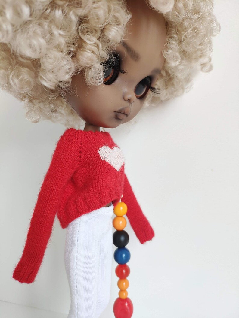 May include: A doll with brown skin and blonde curly hair is wearing a red sweater with a white heart on the front and white pants. The doll is wearing a colorful beaded necklace.