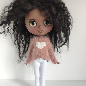 May include: A doll with dark brown curly hair, wearing a pink fuzzy sweater with a white heart, white trousers, and purple and white trainers.