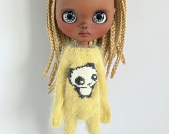 Op de afbeelding: Een pop met een donkere huid en grote blauwe ogen, gekleed in een gele fuzzy romper met een zwart-wit schedelontwerp. De pop heeft blond gevlochten haar en lichtblauwe schoenen.