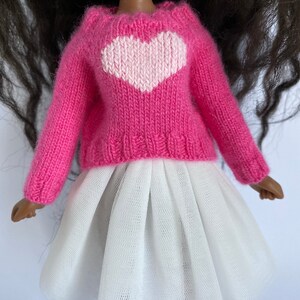 May include: A doll wearing a pink knitted sweater with a white heart design and a white tulle skirt. The doll has dark hair and brown skin. The sweater has long sleeves.