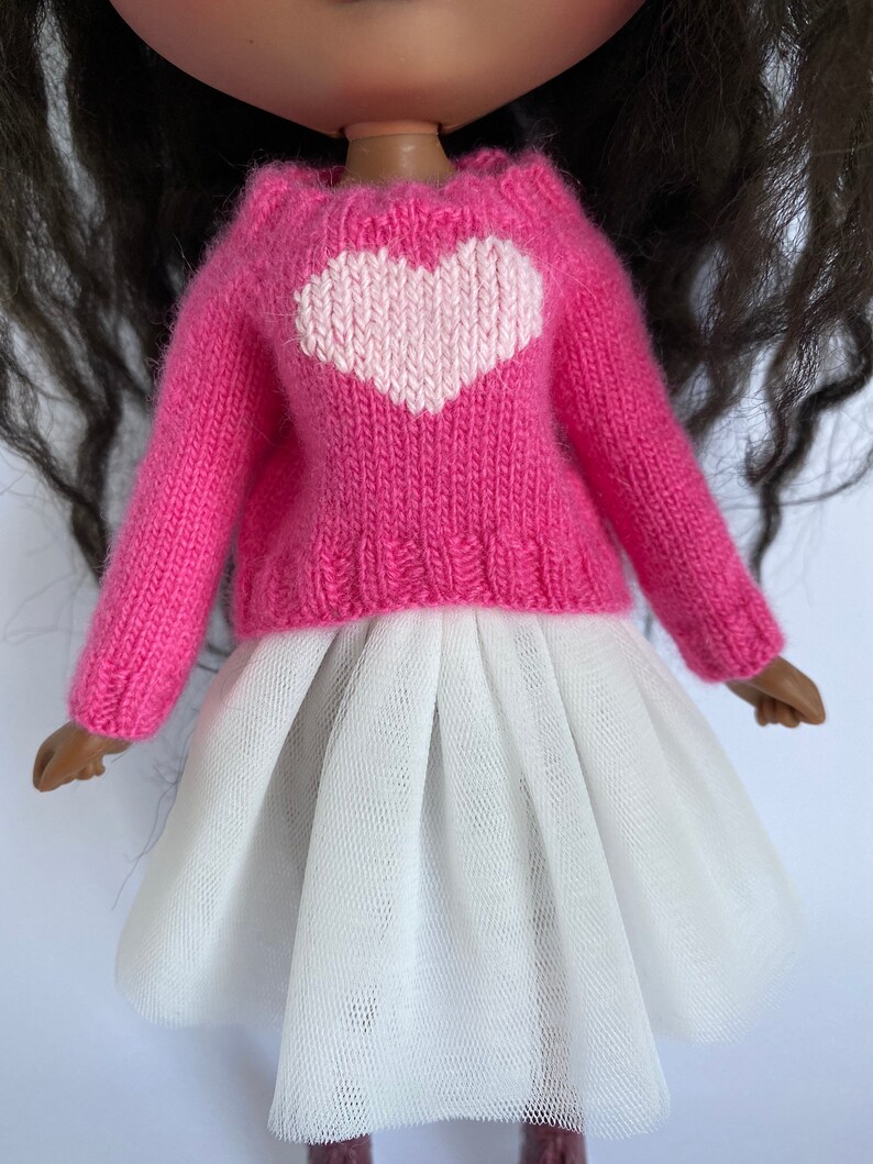 May include: A doll wearing a pink knitted sweater with a white heart design and a white tulle skirt. The doll has dark hair and brown skin. The sweater has long sleeves.