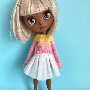 May include: A doll with blonde hair and brown skin wearing a pink and yellow striped sweater and a white tulle skirt. The doll is wearing brown shoes.