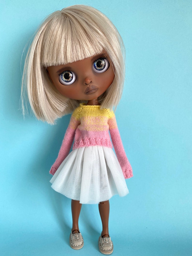 May include: A doll with blonde hair and brown skin wearing a pink and yellow striped sweater and a white tulle skirt. The doll is wearing brown shoes.