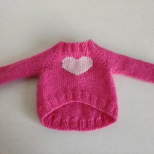May include: A bright pink, hand-knitted sweater with a white heart design on the front. The sweater has long sleeves and a ribbed neckline and hem. This cozy garment is likely for a small pet.