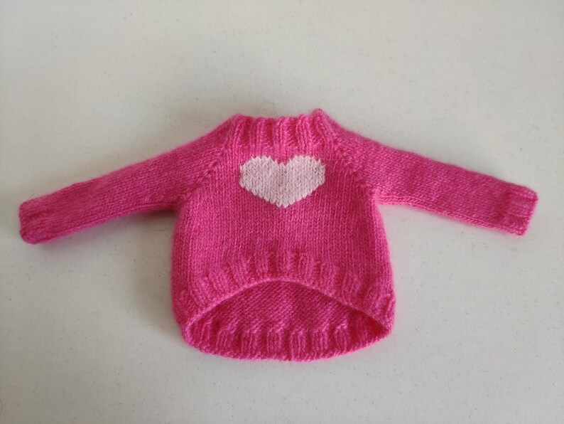 May include: A bright pink, hand-knitted sweater with a white heart design on the front. The sweater has long sleeves and a ribbed neckline and hem. This cozy garment is likely for a small pet.