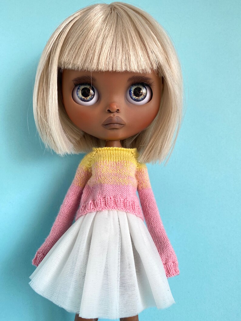 May include: A doll with blonde hair and a pink and yellow knitted sweater, wearing a white tulle skirt.