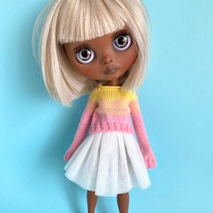 May include: A doll with blonde hair and a bob haircut wearing a pink and yellow striped sweater and a white tulle skirt. The doll has brown skin and blue eyes.