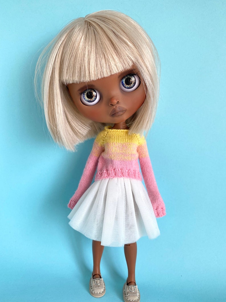 May include: A doll with blonde hair and a bob haircut wearing a pink and yellow striped sweater and a white tulle skirt. The doll has brown skin and blue eyes.