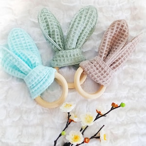 May include: Three baby teething toys in pastel colors. Each toy features a wooden ring and waffle-textured fabric shaped like bunny ears. The colors are light blue, sage green, and a muted pink. A sprig of white flowers with orange accents is in the foreground.