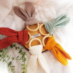May include: Set of four wooden teethers with bunny ear fabric covers. The teethers are in the colors of beige, green, orange, and red. The fabric is a waffle weave texture. The teethers are arranged in a circle on a wooden surface. The text "baby-totti sewing shop" is visible on the wooden surface.
