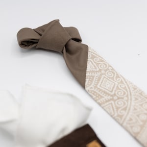 Può includere: Una cravatta con una sezione superiore marrone tinta unita e una sezione inferiore con un motivo geometrico beige e bianco. Accompagnano la cravatta un fazzoletto da taschino bianco e un accessorio in tessuto marrone scuro con una piccola etichetta.
