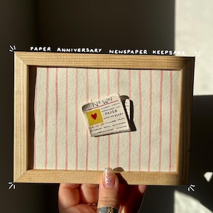 May include: A wooden framed paper anniversary keepsake. The background has vertical pink and white stripes. A small newspaper cutout with the words "NEW!" and "PAPER ANNIVERSARY" is attached. The newspaper has a heart and the date 04.10.24.