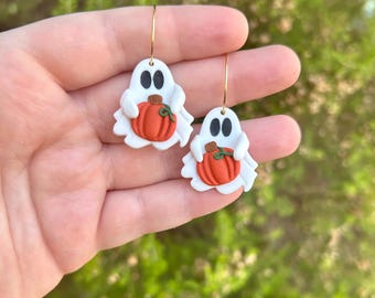 May include: A pair of Halloween-themed earrings. Each earring features a white ghost holding an orange pumpkin. The ghosts have black oval eyes and are attached to gold-colored hoop earrings. The background is a blurred green.
