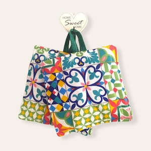 May include: Set of three colourful quilted pot holders with a geometric tile pattern. The pot holders are green, blue, yellow, red and white. The pot holders are hanging from a hook with a heart-shaped sign that says "Home Sweet Home".