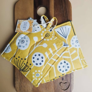 May include: Two yellow pot holders with a white and black floral pattern. The pot holders are on a wooden chopping board with a heart-shaped cutout. The pot holders have a tag that says "Ovenly".