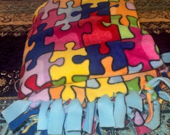 May include: A colorful fleece blanket featuring a jigsaw puzzle pattern in various colors, including red, blue, yellow, and green. The blanket has a light blue fringe along the edge. The blanket is folded on a patterned bedspread.