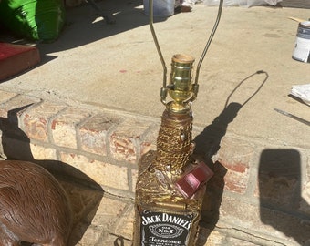 May include: A gold-painted Jack Daniel's whiskey bottle lamp with a gold-coloured lampshade. The bottle is decorated with gold chains and other embellishments. The lamp is sitting on a brick surface.