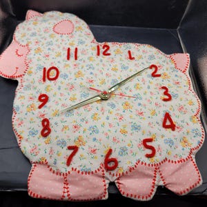 May include: A fabric wall clock in the shape of a sheep, featuring a floral design. The clock has red numerals and hands, with pink detailing. The design is charming and decorative.