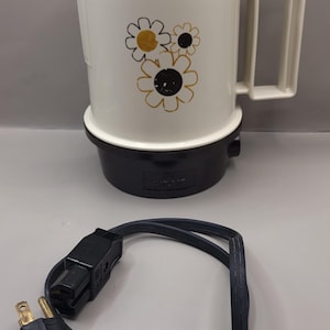 May include: Cream-coloured electric coffee pot with a black base and handle. The pot features a floral design in black and gold. A black power cord with a two-pin plug is coiled below the pot.