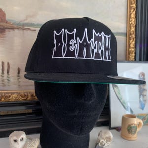 May include: Black baseball cap with a flat brim and white embroidered text. The cap features a green underside and is displayed on a black mannequin head. The text on the cap reads "MEMATH".