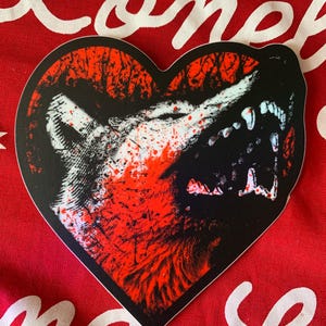 May include: Heart-shaped sticker featuring a snarling wolf's head in black, red, and white. The design includes splatters of red, creating a dramatic and intense visual. The sticker is set against a red background.