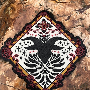 May include: A black and white embroidered patch with a stylized bird design. The bird has large wings and a small head. The patch is surrounded by a red and gold border with floral details.
