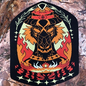 Puede incluir: Un parche bordado negro con un diseño de dragón naranja y amarillo fuego. El dragón tiene un tercer ojo y está rodeado de llamas y estrellas. El texto "Rise" está escrito en una fuente estilizada en la parte inferior del parche.