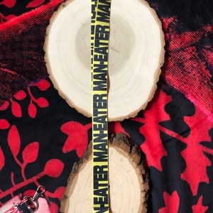 May include: A yellow lanyard with the words "MAN EATER" in black, repeated vertically. The lanyard has a silver-colored metal clasp. The background includes a wooden slice and a black and red patterned fabric.