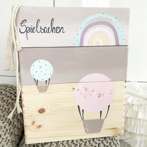 Könnte beinhalten: Eine dreistufige Holzaufbewahrungsbox, die in sanften Rosa- und Hellbrauntönen lackiert ist. Die oberste Stufe zeigt einen Regenbogen mit goldenen Glitzerakzenten und das Wort "Spielachen" in schwarzer Schrift. Die mittlere Stufe hat einen Heißluftballon mit goldenen Punkten. Die unterste Stufe hat einen Heißluftballon mit rosa und weißen Blumenakzenten.