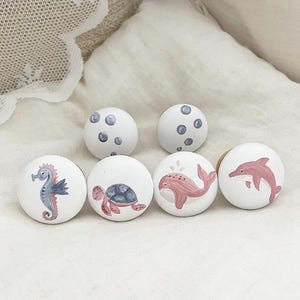 Könnte beinhalten: Sechs runde Holzgriffe mit bemalten Meeresmotiven. Die Griffe zeigen ein Seepferdchen, eine Schildkröte, einen Wal, einen Delfin und zwei Griffe mit blauen und weißen Punkten.