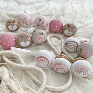 Könnte beinhalten: Ein Set mit 11 Holz-Schubladengriffen mit verschiedenen Designs. Die Griffe sind in Rosa, Weiß und Braun lackiert und mit Motiven von Hirschen, Blumen und Regenbögen verziert.