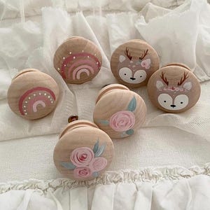 Könnte beinhalten: Sechs Holzgriffe mit verschiedenen bemalten Designs. Die Designs beinhalten einen Regenbogen, Blumen und ein Reh. Die Griffe sind alle in zarten Rosa-, Weiß- und Brauntönen bemalt.
