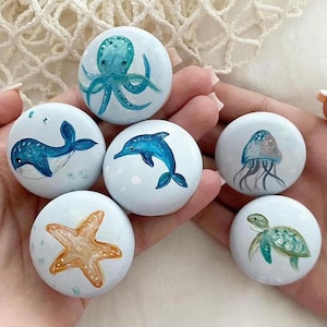 May include: Six painted round stones with different ocean-themed designs. The stones are painted with a light blue background and feature a blue whale, a dolphin, an octopus, a jellyfish, a starfish, and a sea turtle.