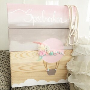 Könnte beinhalten: Eine hölzerne Spielzeugkiste mit einem rosa, weißen und braunen Ombre-Farbdesign. Die Kiste hat einen Heißluftballon mit einem Blumendesign auf der Vorderseite und das Wort "Spielachen" auf der Oberseite.