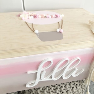 Könnte beinhalten: Eine Holzkiste mit einem rosa-weißen Ombre-Finish. Der Deckel zeigt ein Heißluftballon-Design mit rosa und weißen Blumen und Pompons. Die Box ist mit dem Namen "Lila" in weißen Buchstaben personalisiert.
