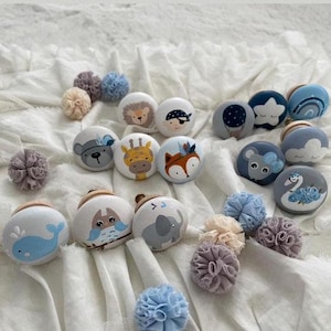 Könnte beinhalten: Eine Sammlung von 14 bemalten Holzknöpfen mit verschiedenen Tiermotiven, darunter ein Wal, eine Eule, ein Elefant, ein Fuchs, eine Giraffe, ein Bär und ein Schwan. Die Knöpfe sind in Blautönen, Grau und Weiß bemalt, einige mit gelben und braunen Akzenten.