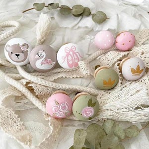May include: A set of nine decorative knobs with various designs, including a bear, a swan, ballet shoes, flowers, and a crown. The knobs are painted in shades of pink, green, and white.