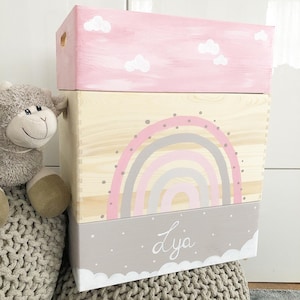 Könnte beinhalten: Drei übereinander gestapelte Holzkisten mit rosa, naturbelassenem Holz und grauer Oberfläche. Die mittlere Kiste zeigt ein Regenbogen-Design mit rosa, grauen und weißen Streifen. Die obere Kiste hat einen rosa Hintergrund mit weißen Wolken. Die untere Kiste hat einen grauen Hintergrund mit weißen Wolken und den Namen "Lya" in weiß.
