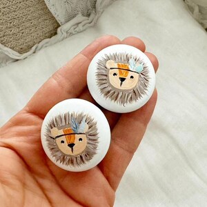 Könnte beinhalten: Zwei weiße runde Holzknöpfe mit bemalten Löwenköpfen. Jeder Löwe hat eine braune Mähne, orange und braune Streifen und blaue und weiße Federn auf dem Kopf.