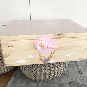 Könnte beinhalten: Eine Holzkiste mit einem hellrosa Deckel. Der Deckel ist mit einem Heißluftballon-Motiv verziert, das mit rosa und weißen Blumen, Pompons und dem Namen "Charlotte" in weißer Schrift verziert ist.