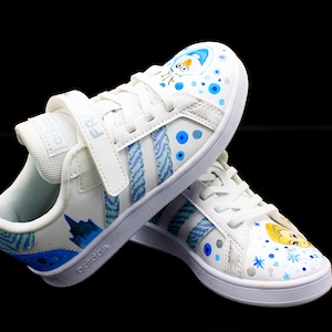 K&ouml;nnte beinhalten: Wei&szlig;e Canvas-Sneaker mit blauen und gr&uuml;nen Streifen und einem blauen und wei&szlig;en Schneeflocken-Design. Die Sneaker haben einen Klettverschluss und das Wort "adidas" ist seitlich aufgedruckt.