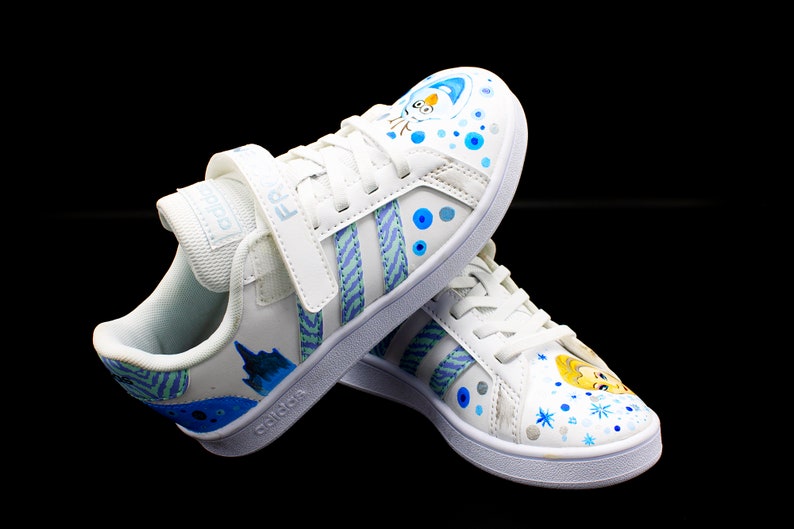 K&ouml;nnte beinhalten: Wei&szlig;e Canvas-Sneaker mit blauen und gr&uuml;nen Streifen und einem blauen und wei&szlig;en Schneeflocken-Design. Die Sneaker haben einen Klettverschluss und das Wort "adidas" ist seitlich aufgedruckt.